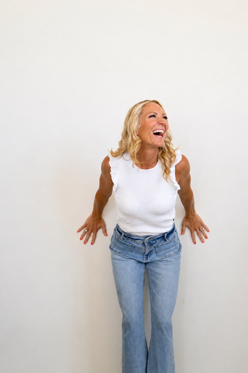 Amy Sanders laughing, leaning back against a white wall in a white top and jeans